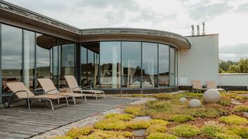A modern building with large glass windows, outdoor seating with lounge chairs, and a landscaped garden area with moss and stones.