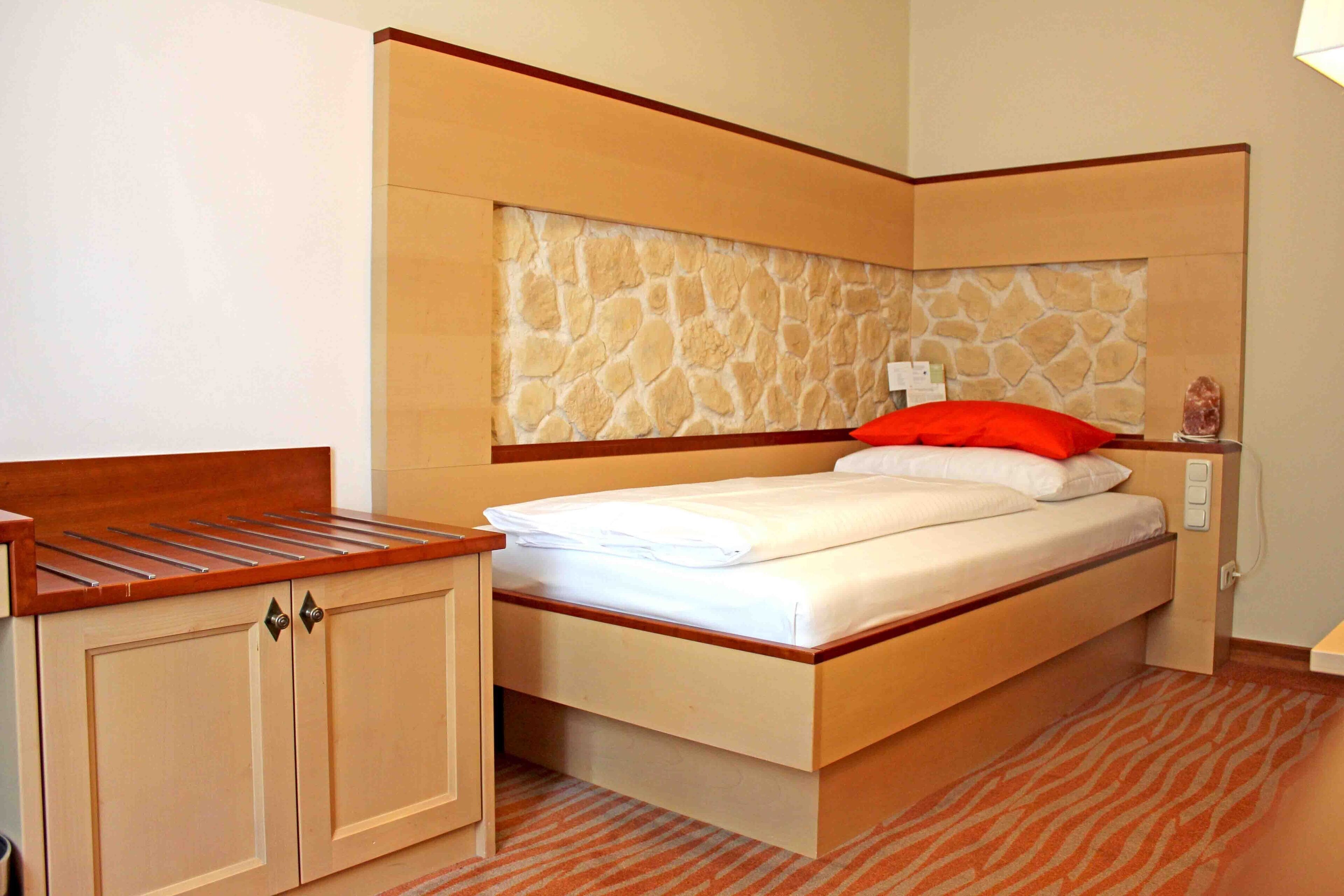 Una habitación de hotel pequeña con una cama, un banco de madera y un armario.
