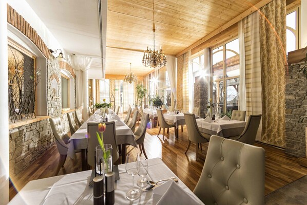 A restaurant with wooden ceilings, stone walls, and large windows. Tables are set with white tablecloths and chairs.