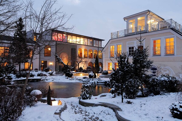 Une maison moderne avec un grand étang, un jardin enneigé et des fenêtres illuminées.