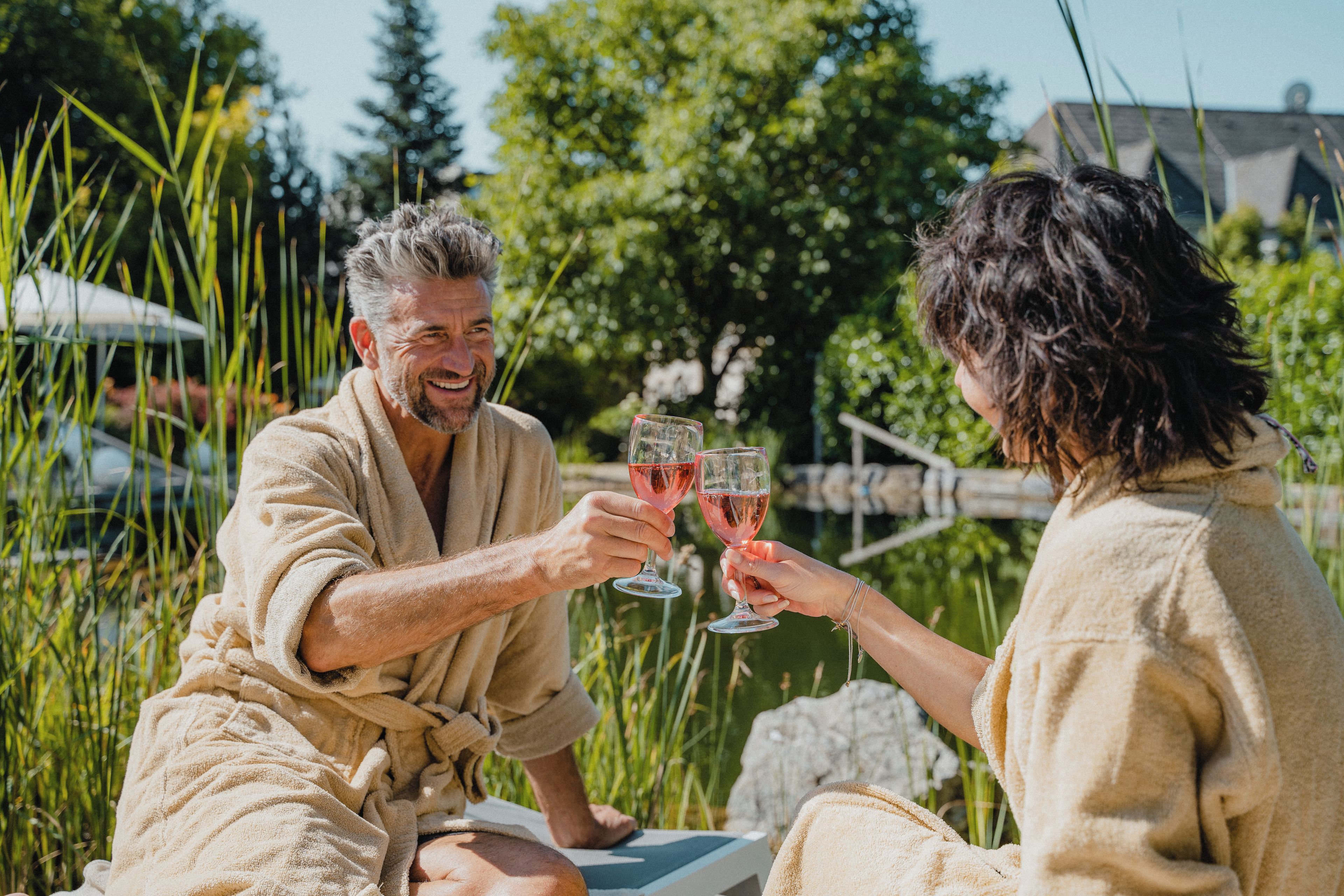 Two people in bathrobes sitting on a lounge chair outdoors, holding wine glasses.