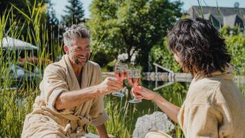 Two people in bathrobes sitting on a lounge chair outdoors, holding wine glasses.