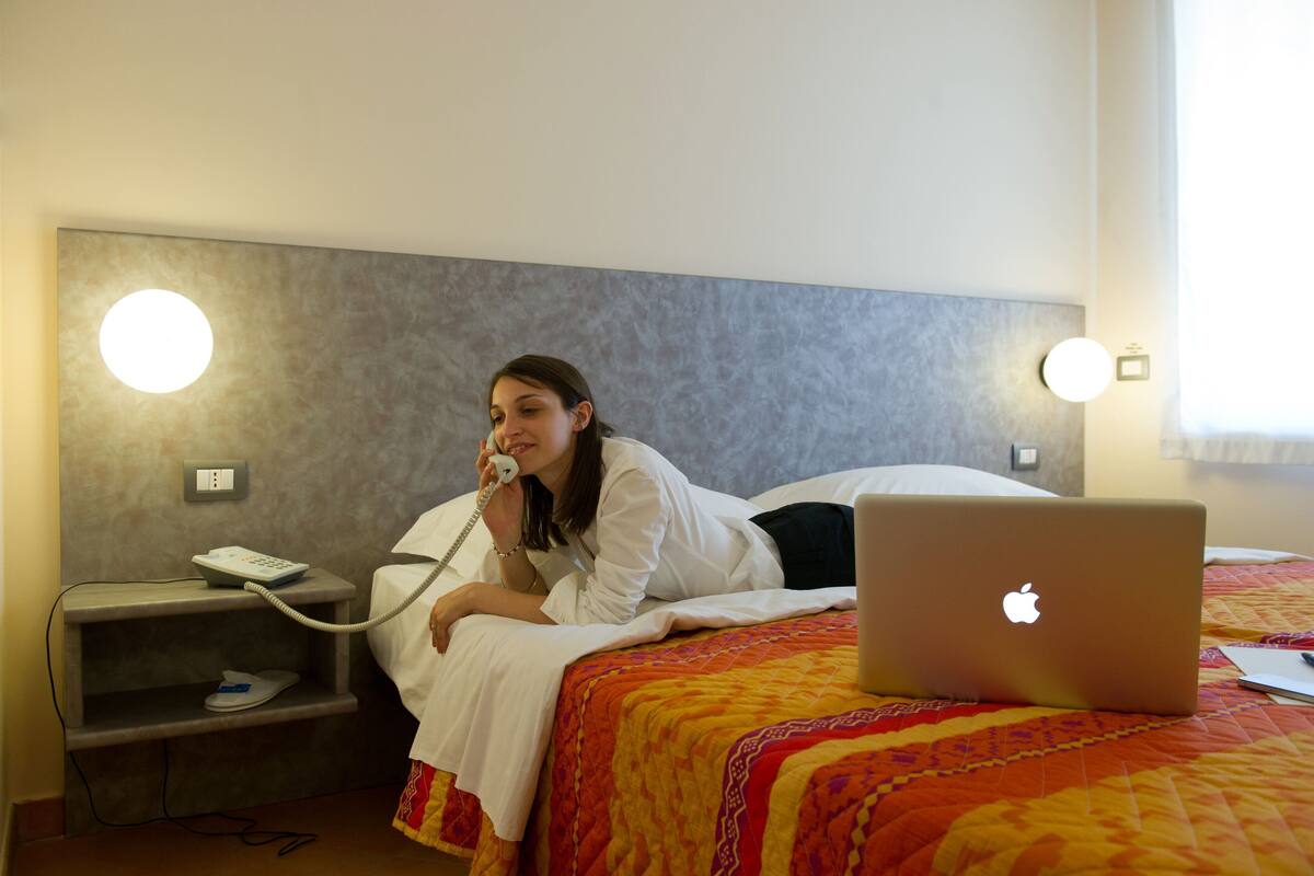 A hotel room with a bed, a bedside table with a phone, and a laptop on the bed.