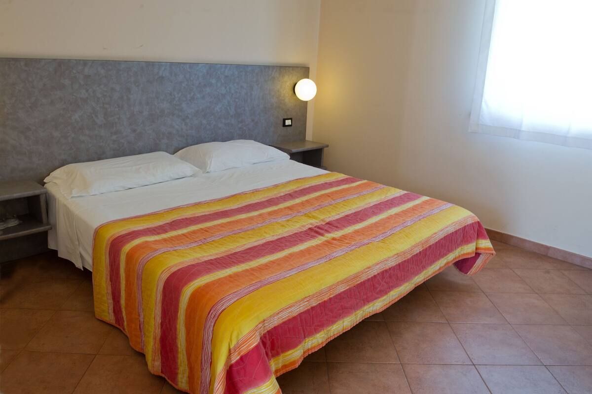 A bed with a striped blanket, a bedside table with a lamp, and a window with curtains.