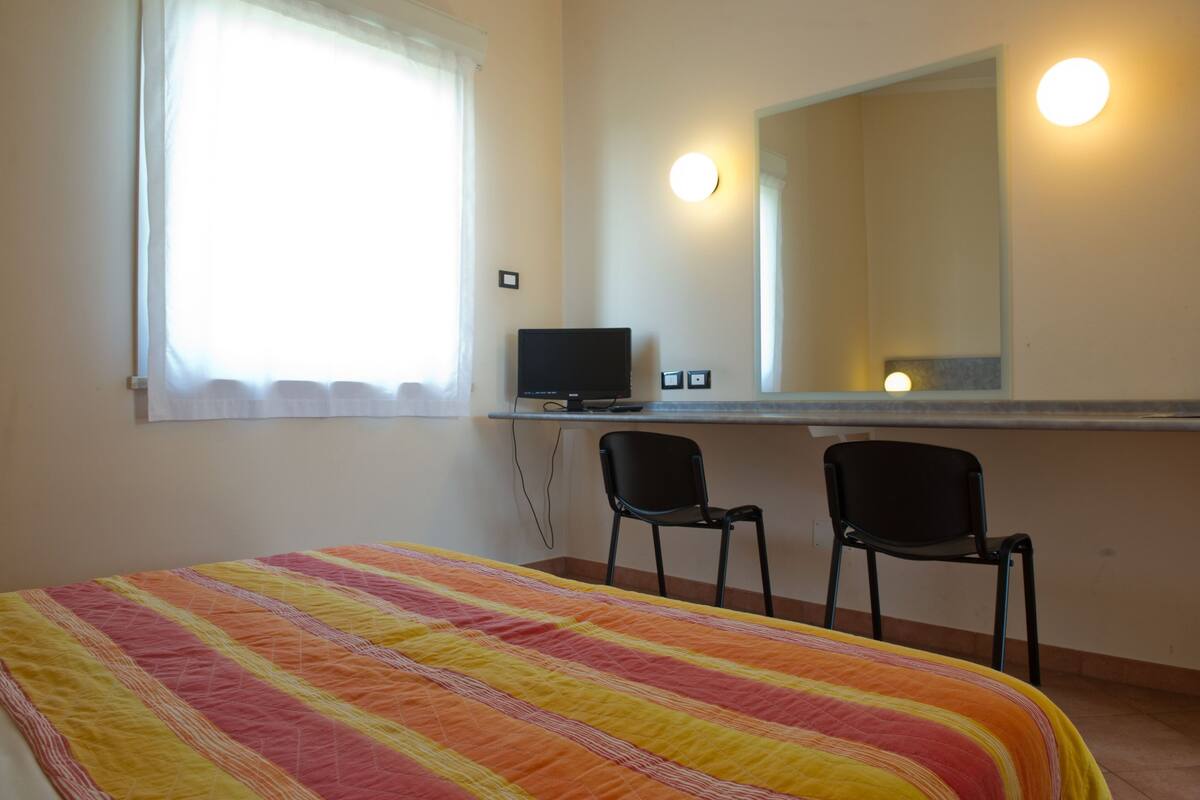 A room with a bed, two chairs, a desk with a computer, a window with curtains, and a mirror with lights.