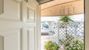 A hotel entrance with a white door marked "115", a covered outdoor area with a lattice fence, and a parked van in the background. - Borgo del Mare (Civitavecchia)