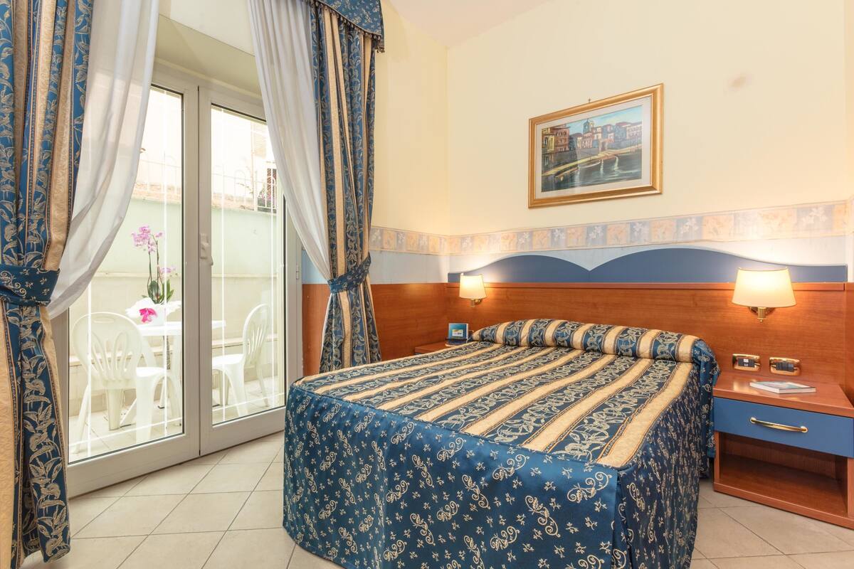 A hotel room with a bed, two bedside tables, a window with curtains, a picture on the wall, and a view of the outside.