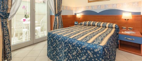 A hotel room with a bed, two bedside tables, a window with curtains, a picture on the wall, and a view of the outside.