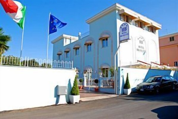 A hotel with a white facade, a signboard with 'HB' and 'Hotel', and two flags on poles in front.