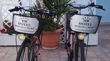 Dos bicicletas con cestas que muestran la leyenda "HB HOTEL BORGO del MARE" estacionadas frente a un edificio con una cerca de celosía blanca y una maceta grande.