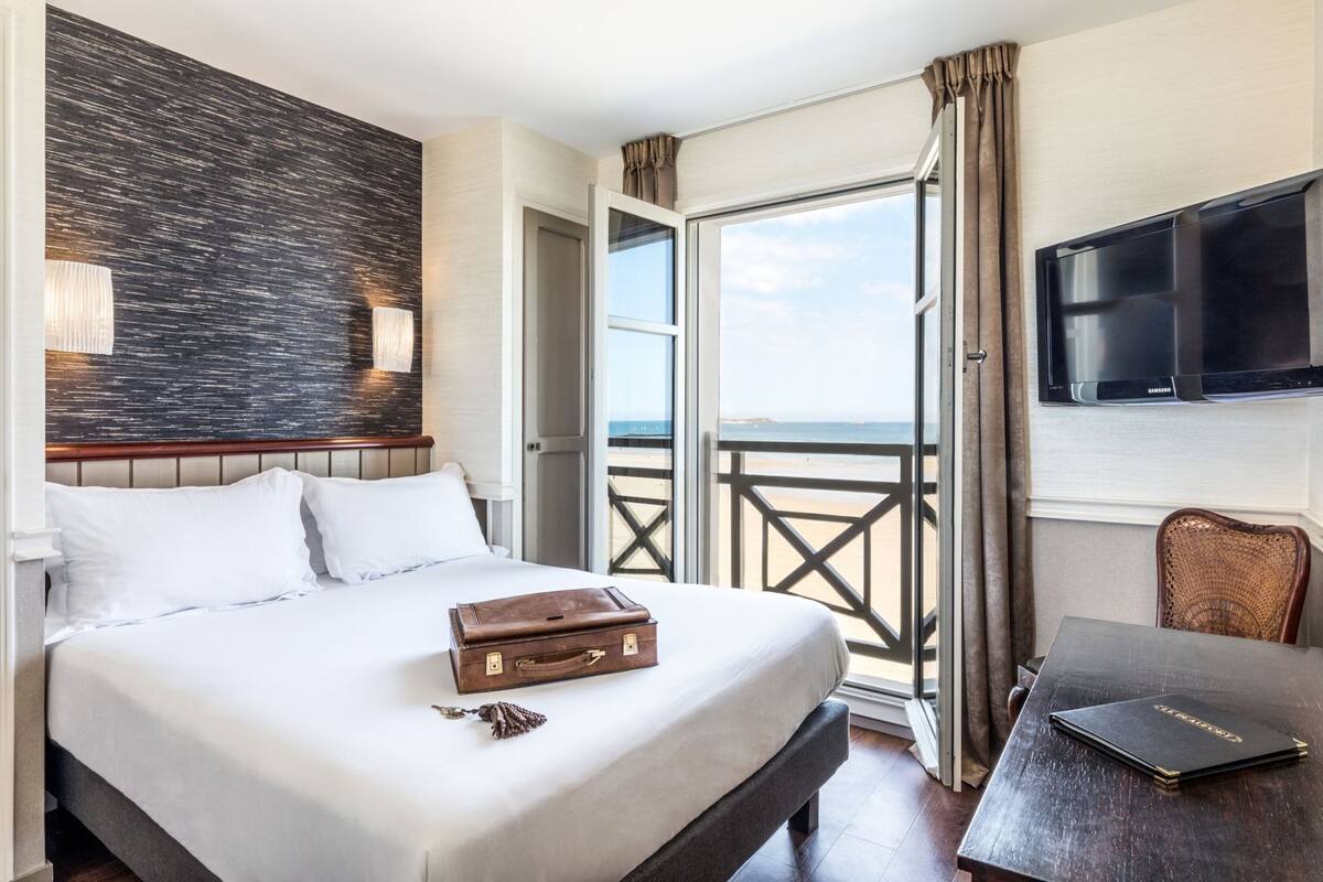 Ein Hotelzimmer mit Bett, Koffer, Schreibtisch mit Laptop, Fernseher und einem Balkon mit Blick.