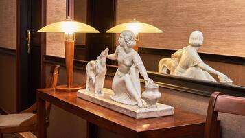 A marble statue of a woman with a child on a wooden table, illuminated by two lamps, with a mirror reflecting the statue.