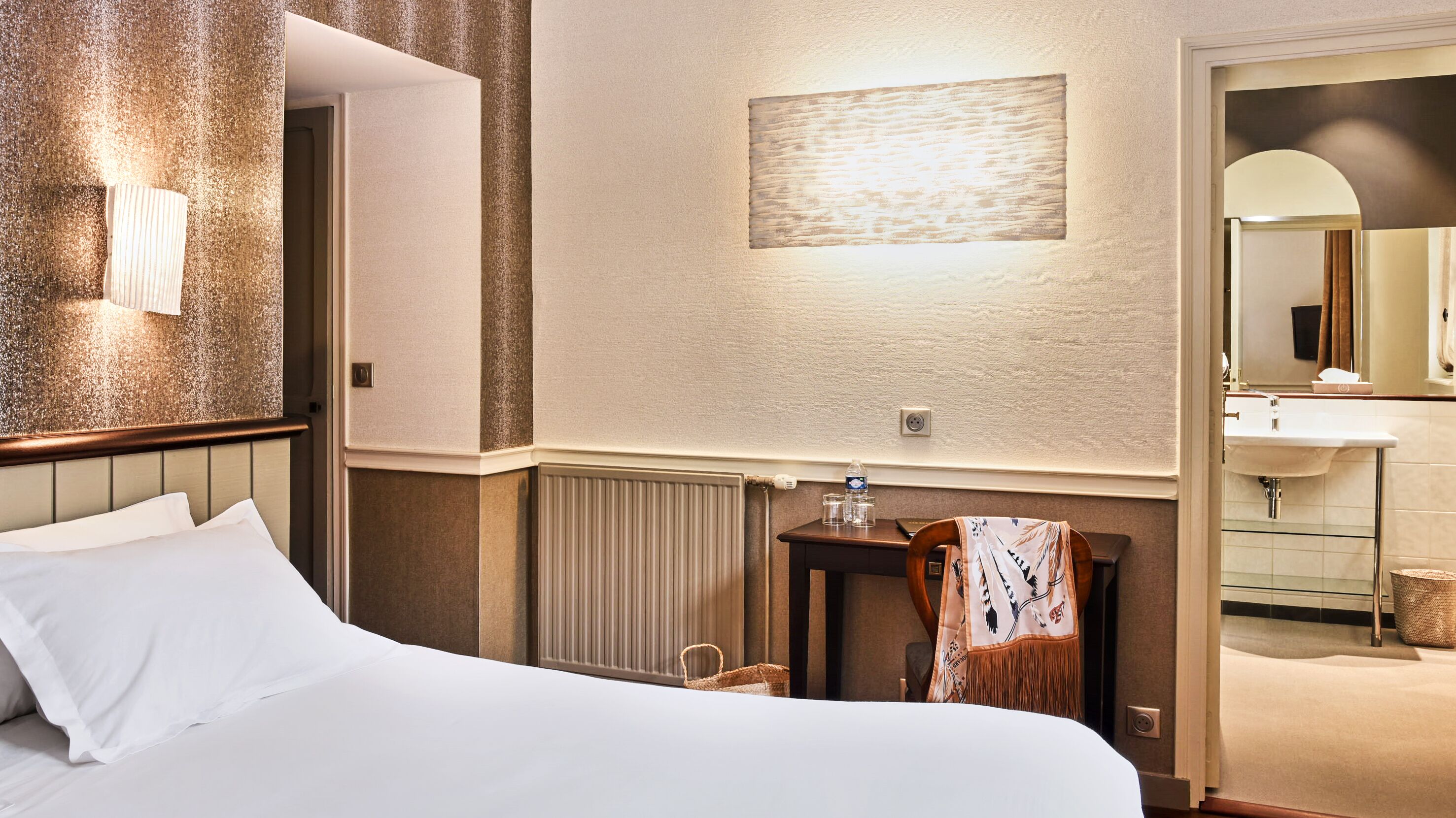 Une chambre d’hôtel avec un lit, une table de chevet sur laquelle se trouvent une bouteille et des verres, une chaise recouverte d’un tissu à motifs, un lampadaire fixé au mur et une salle de bain visible à travers une porte ouverte.
