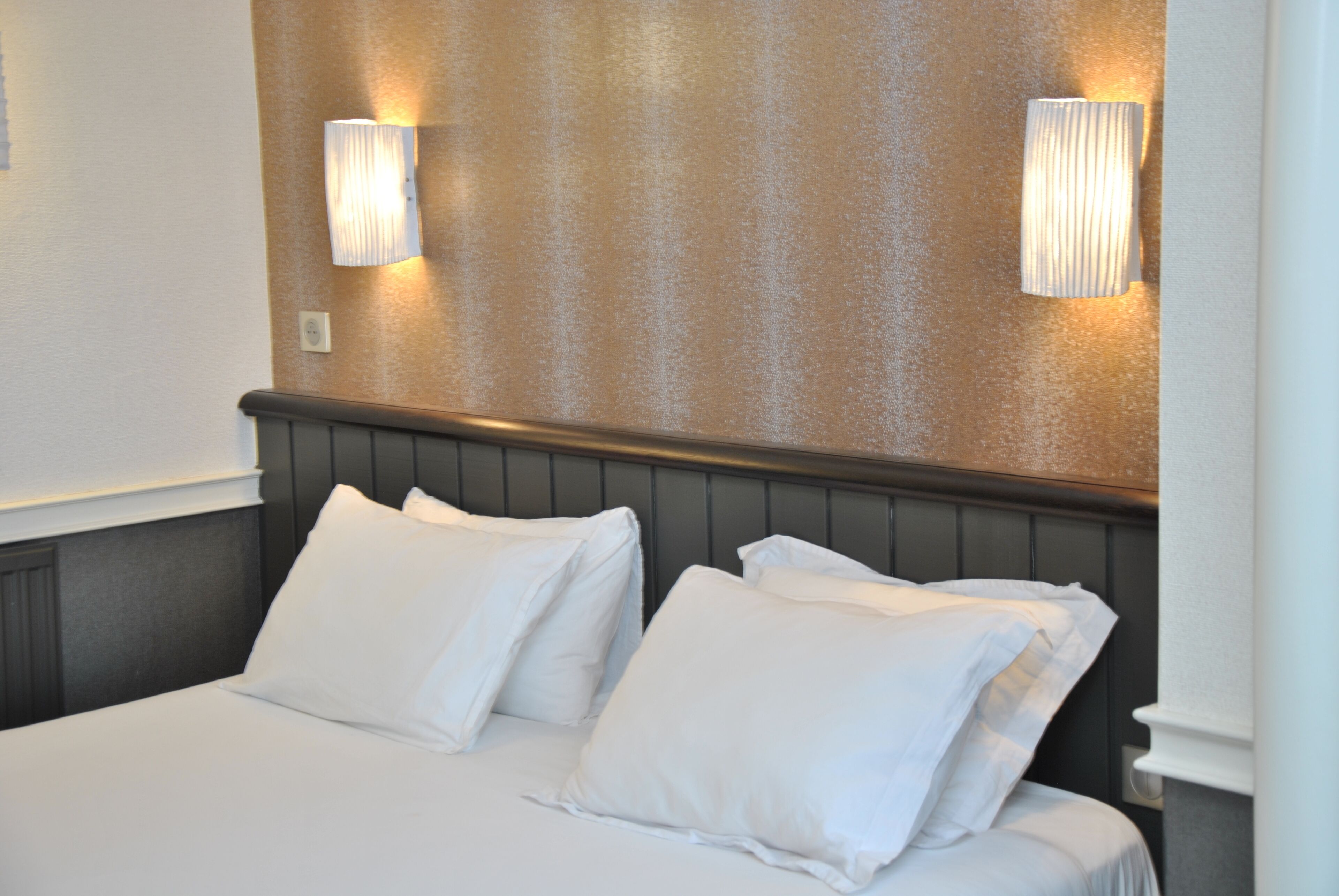 Een net opgemaakt bed met witte lakens en kussens, een houten hoofdbord en twee wandlampen op een structuurwand.
