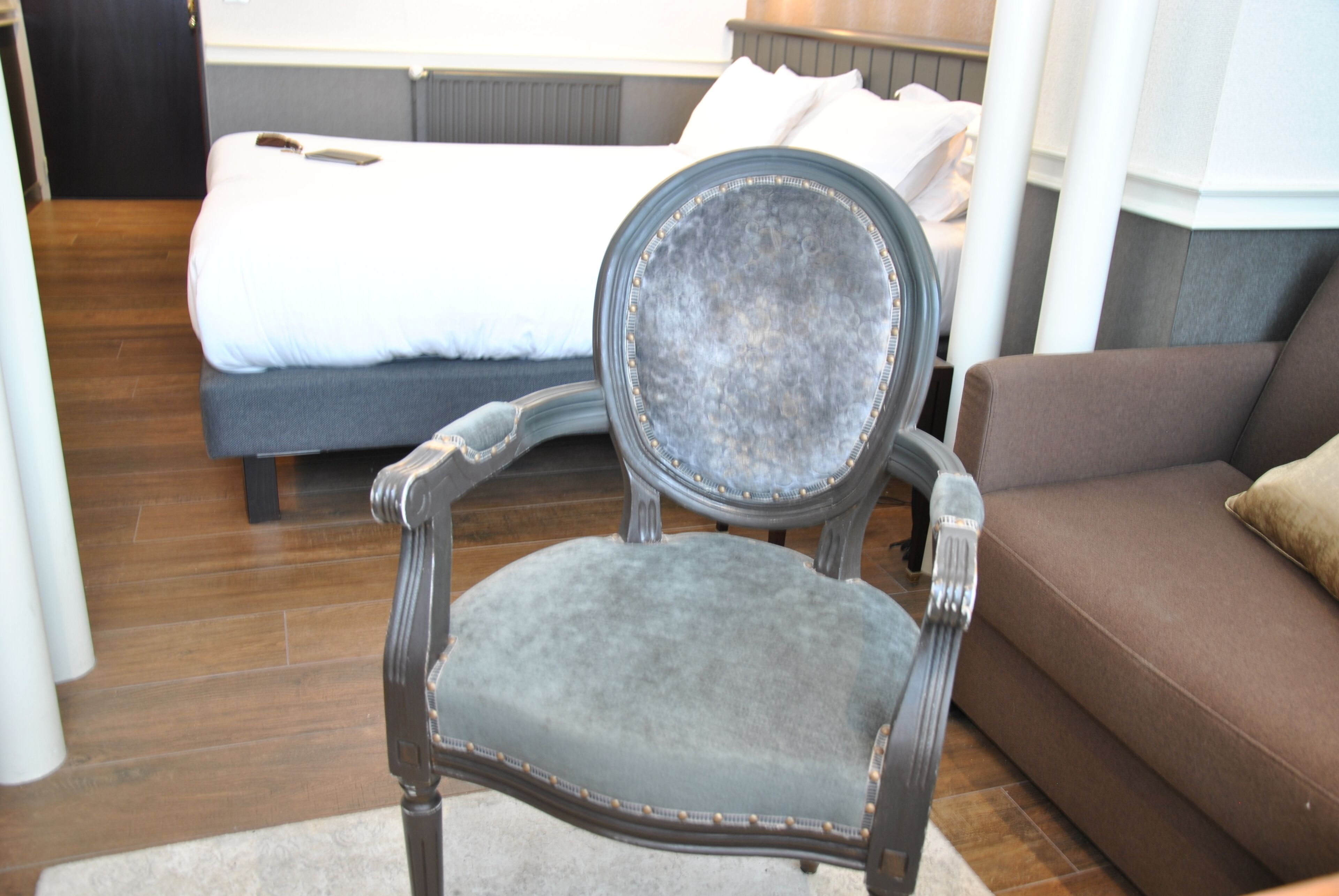 A room with a bed, a brown sofa, and a grey upholstered chair with a metal frame.