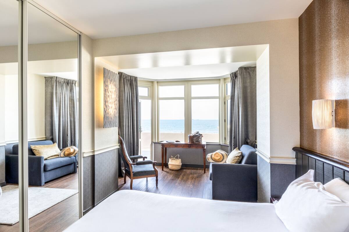 Ein Hotelzimmer mit einem großen Bett, zwei Sesseln, einem kleinen Tisch und Meerblick durch große Fenster.