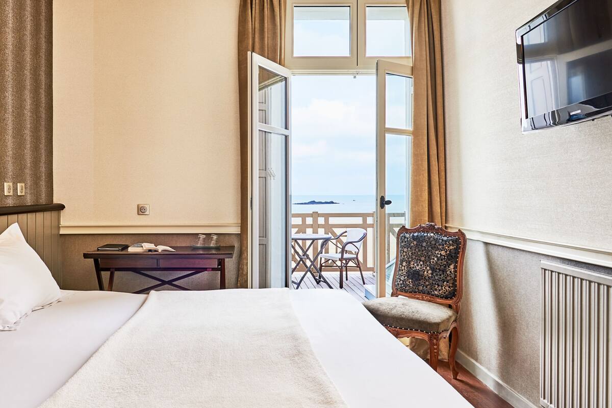 Ein Hotelzimmer mit Bett, Schreibtisch mit Stuhl, Fernseher und einem Balkon mit Meerblick.
