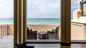 Varanda com cadeiras de vime e uma mesa, com vista para uma praia e águas calmas, com um navio ao longe.