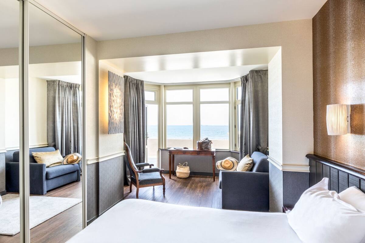Ein Hotelzimmer mit Bett, einer Sitzecke inklusive Sofa und Sesseln, einem kleinen Tisch und Meerblick durch große Fenster.