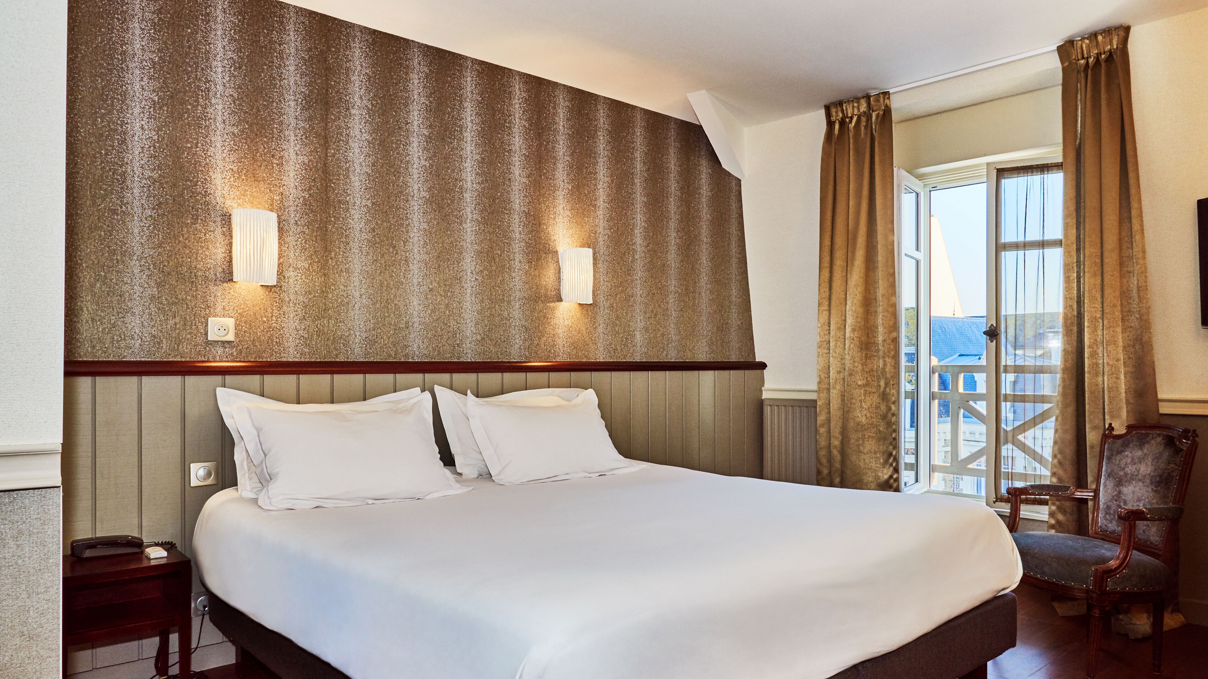 A hotel room with a large bed, a wooden headboard, two wall-mounted lights, a chair, and a view of the outside through a window.