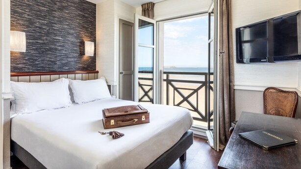Quarto de hotel com uma cama, uma mala, uma mesa com um livro, uma televisão e uma varanda com vista.