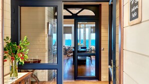 Visão através de uma porta de vidro para uma área de refeições com mesa e cadeiras, vista para o mar e um letreiro indicando um restaurante.