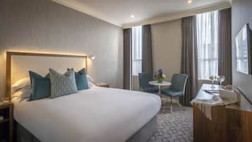 Habitación de hotel con una cama grande, un escritorio con dos sillas, una mesita redonda y un ventanal con cortinas.