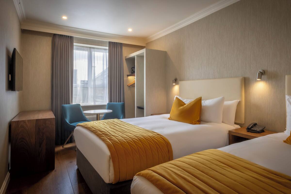 Habitación de hotel con dos camas, un escritorio, una silla y un ventanal con cortinas.