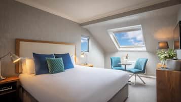 A hotel room with a large bed, a skylight, a small table with chairs, and a nightstand with a lamp.