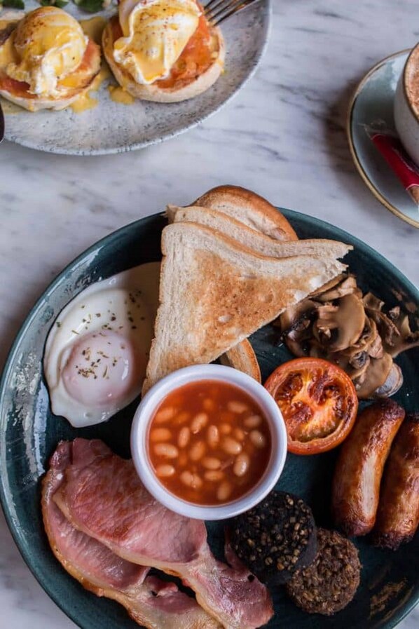 Un plato de desayuno con huevos benedictinos, tostadas, frijoles, tocino, champiñones, salchichas y tomate.