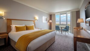 Habitación de hotel con una cama grande, dos lámparas de noche, un escritorio con silla y un ventanal con vista al mar.