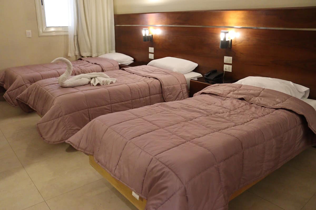 Ein Hotelzimmer mit drei Betten, jedes mit einer rosa Tagesdecke und weißen Kissen. Über den Betten sind Wandleuchten angebracht und ein Telefon steht auf dem Nachttisch.