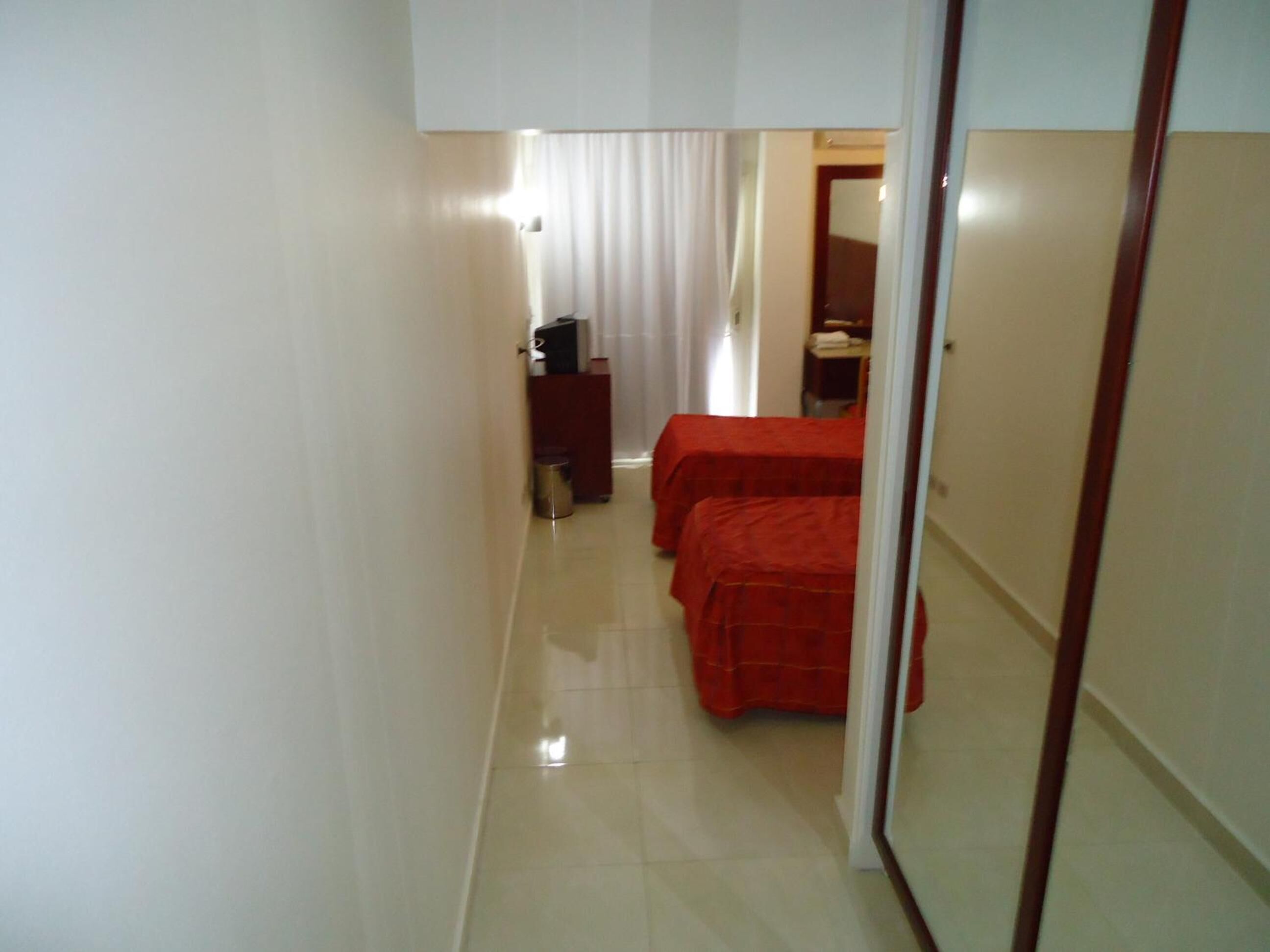 A narrow hallway with two beds covered in red sheets, a small desk with a computer, and a trash can.