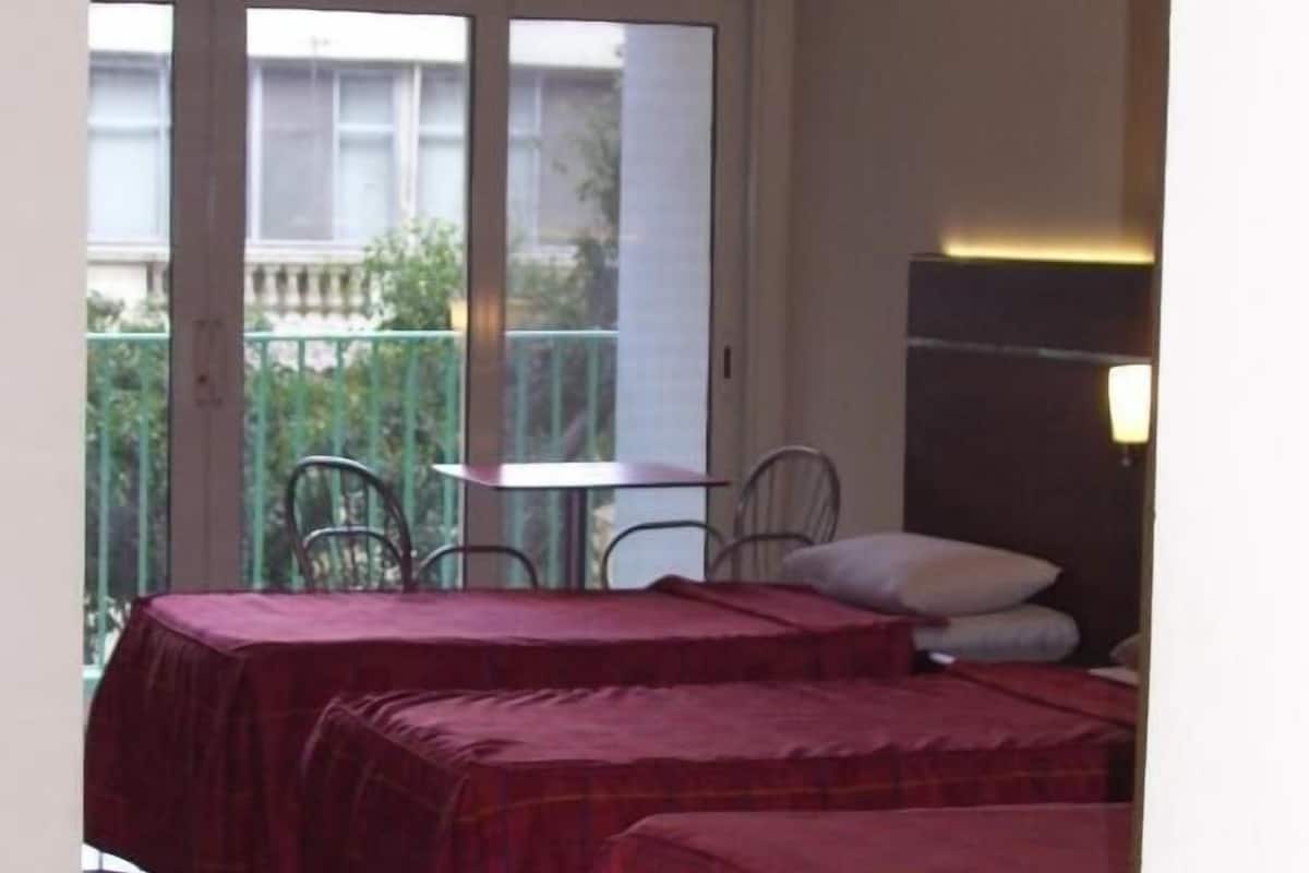 Ein Zimmer mit zwei Betten, einem Tisch und Stühlen, mit Balkon, der durch das Fenster sichtbar ist.