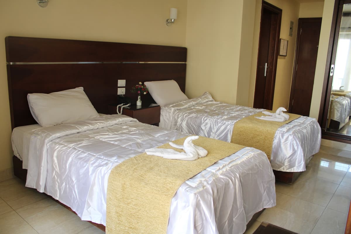 Ein Hotelzimmer mit zwei Betten, jeweils mit weißen Leintüchern und einer beige farbenen Tagesdecke. Ein Nachttisch mit Lampe und einer Blumenvase ist ebenfalls vorhanden.