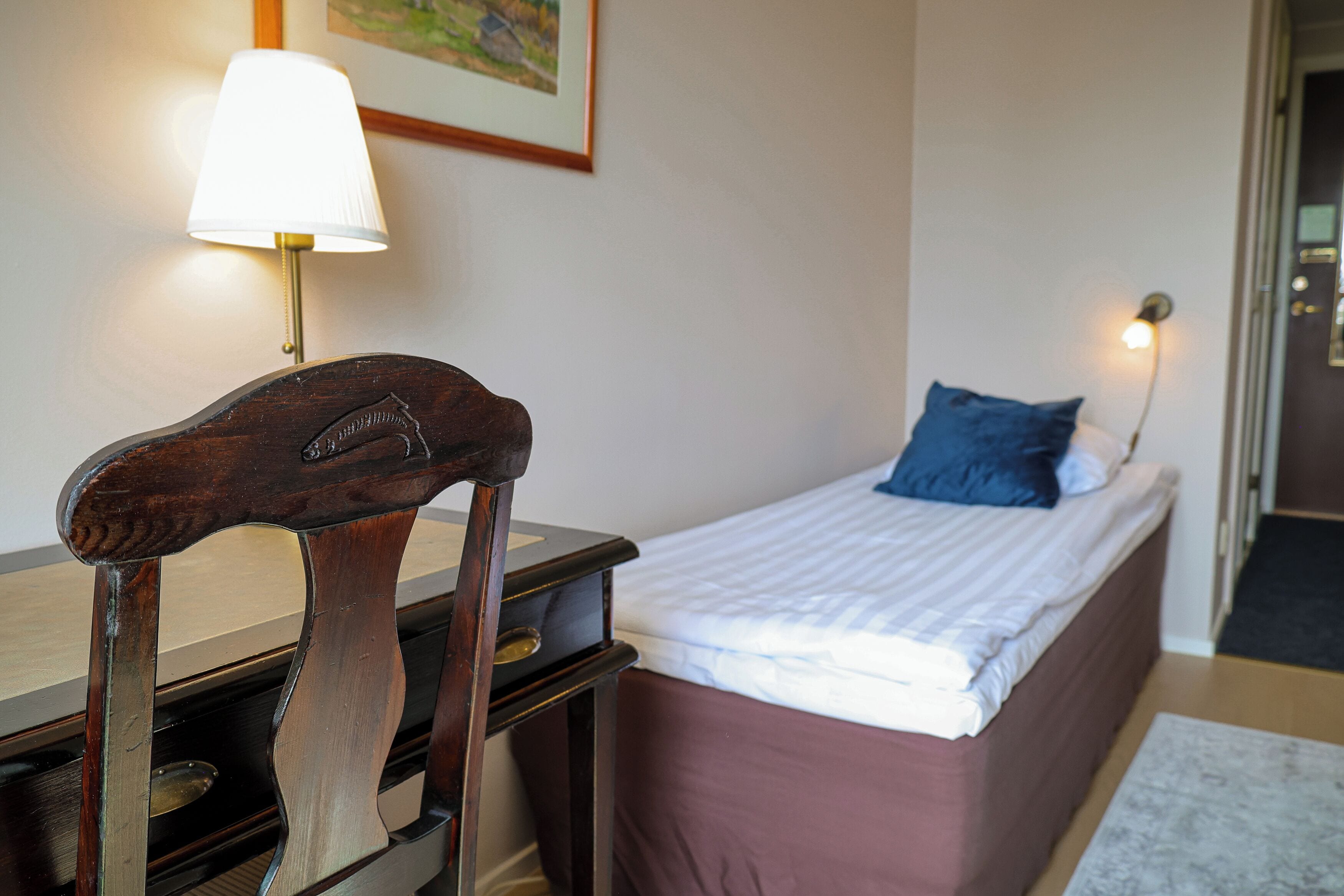 A bedroom with a single bed, a wooden chair, a desk, a lamp, and a framed picture on the wall.
