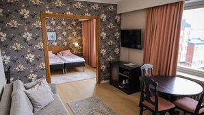 Hotellihuone, jossa on sänky, sohva, ruokapöytä tuoleineen, televisio ja ikkuna, jossa on verhot.