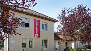 A building with a beige facade, a red 'APART' sign, and a small garden area with a tree. - Appart’City Classic Dijon – Toison D’or (Ahuy)