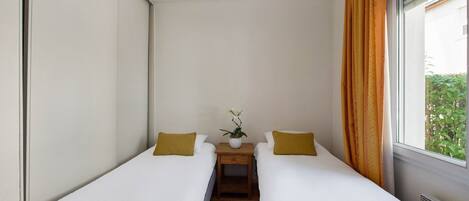 Una habitación pequeña y moderna con dos camas individuales, una mesita con un florero y una ventana con cortinas.