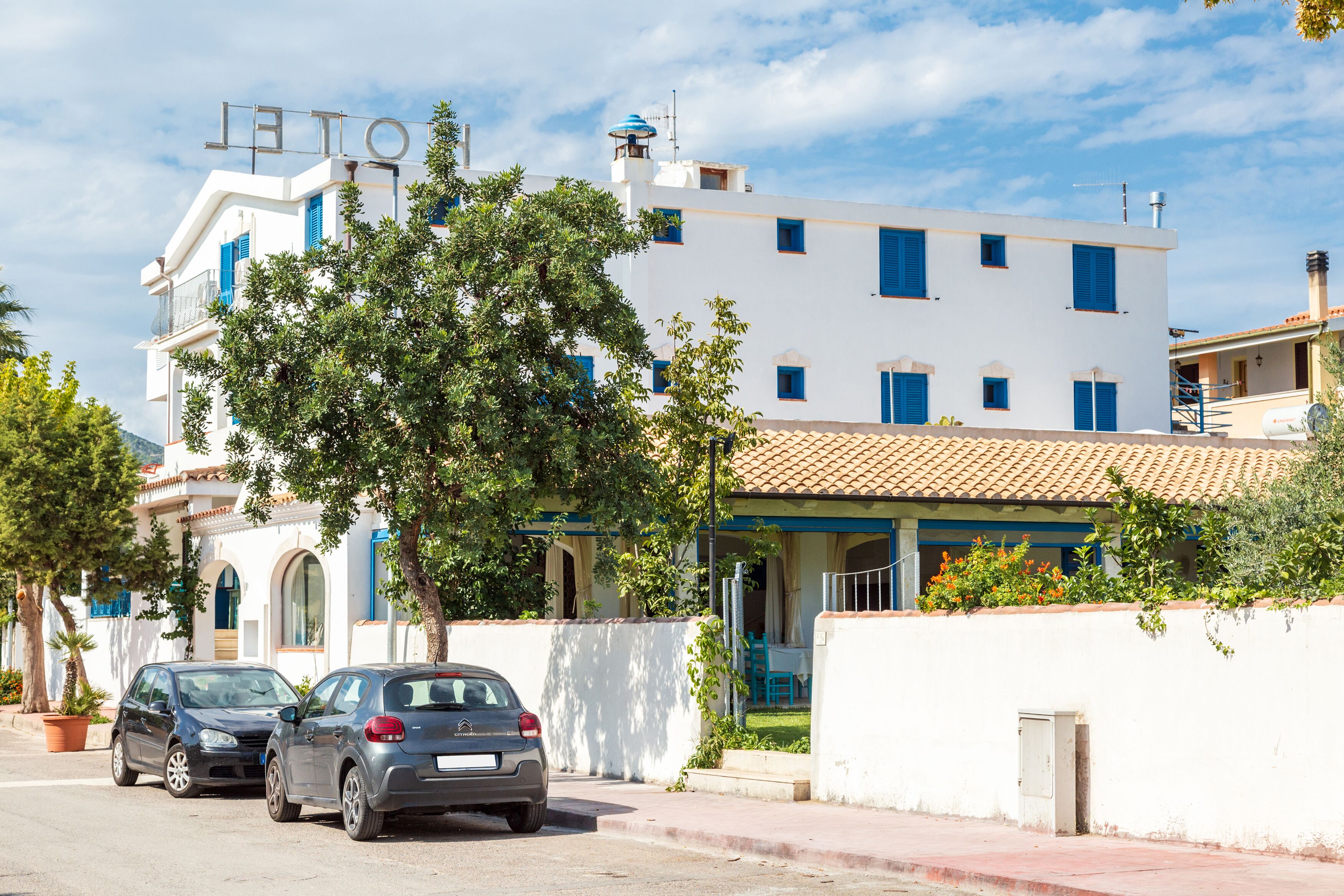 A white building with blue shutters and a tiled roof. There are two cars parked in front.