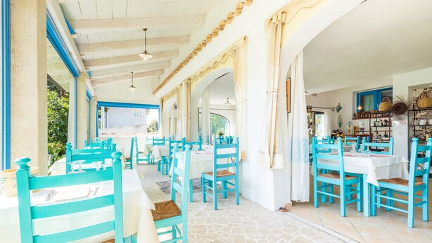 Ein Restaurant mit türkisfarbenen Stühlen und weißen Tischen, mit Steinboden und Blick ins Freie.