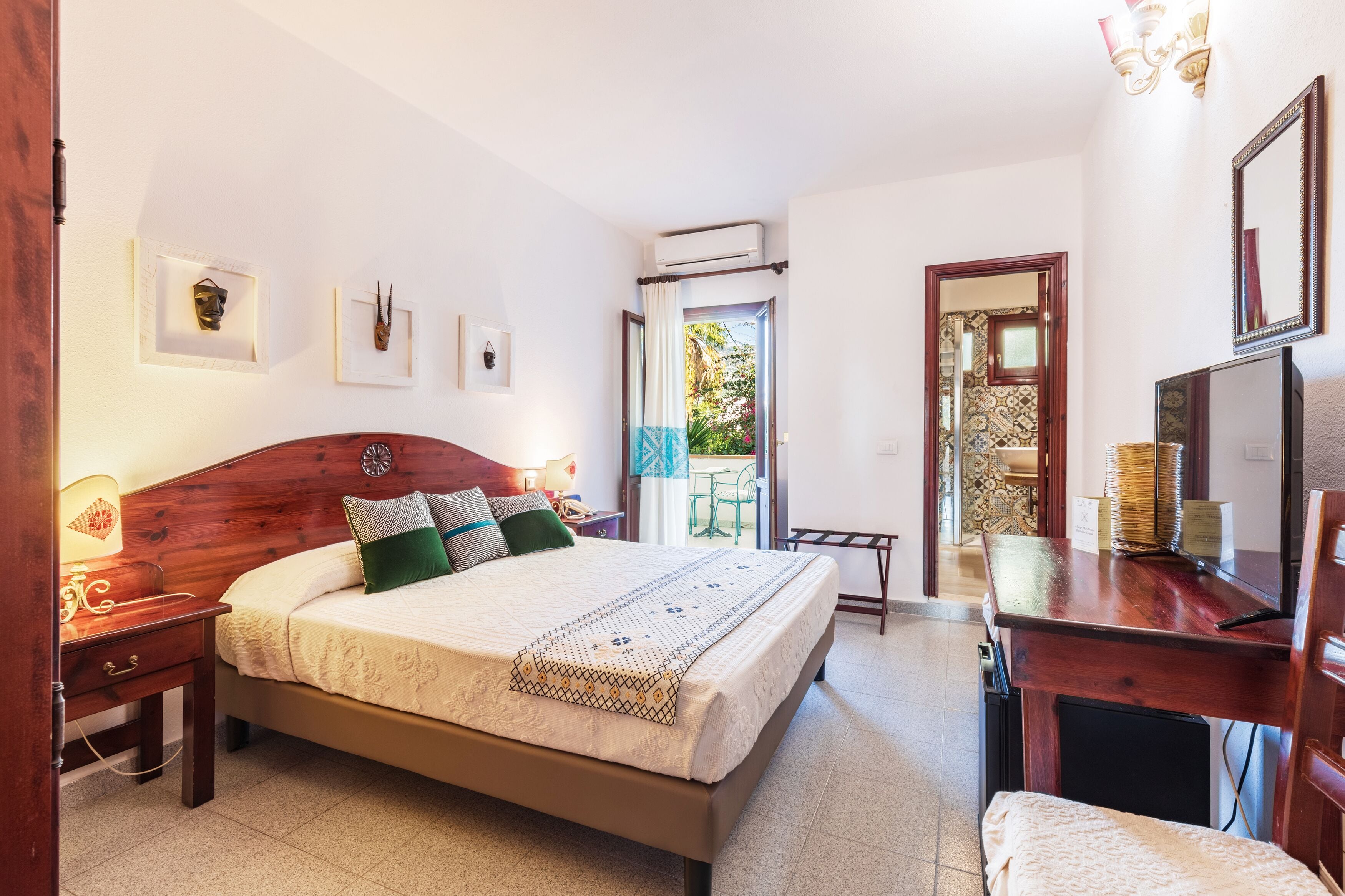 Un dormitorio con una cama de madera, mesitas de noche, un escritorio y una silla. Se ve una ventana con cortinas, una puerta y un aire acondicionado instalado en la pared.