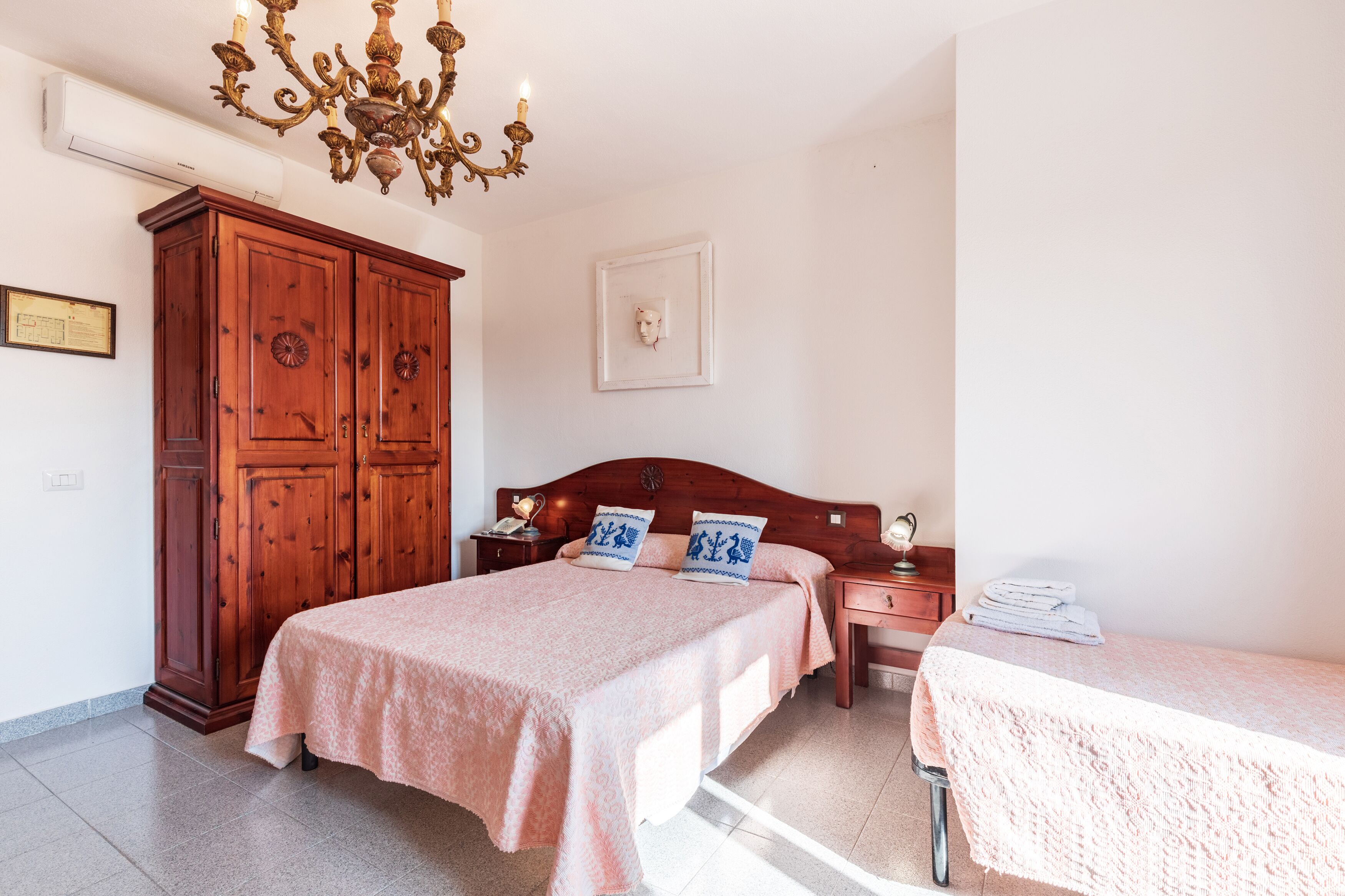 A bedroom with a wooden wardrobe, a bed with a pink bedspread, a bedside table with a lamp, and a small table with towels.