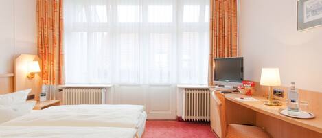 A hotel room with two beds, a desk with a computer, a chair, a TV, and a radiator.
