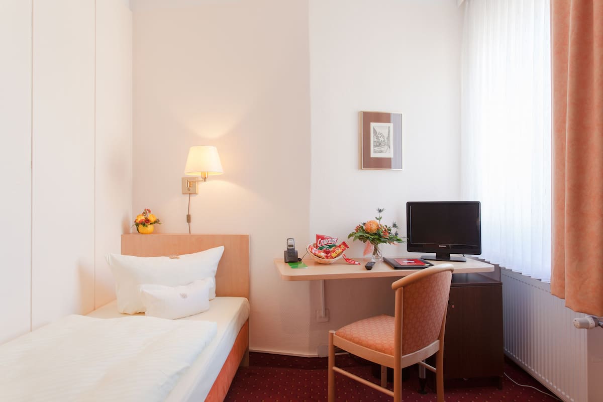 Habitación de hotel con cama, escritorio con computadora, silla, lámpara y ventana con cortinas.