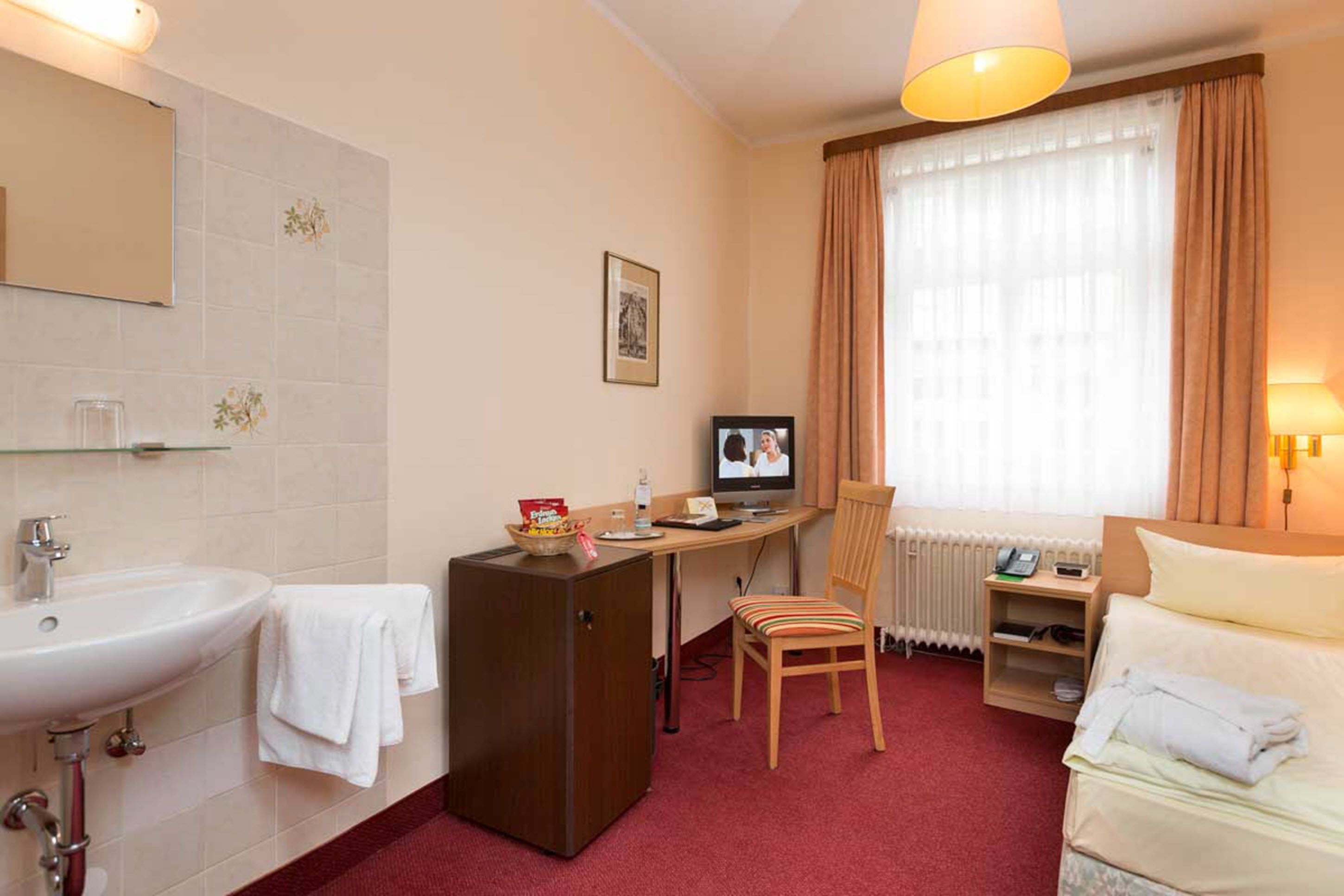 A hotel room with a bed, a desk with a computer, a chair, a small refrigerator, a sink, and a window with curtains.