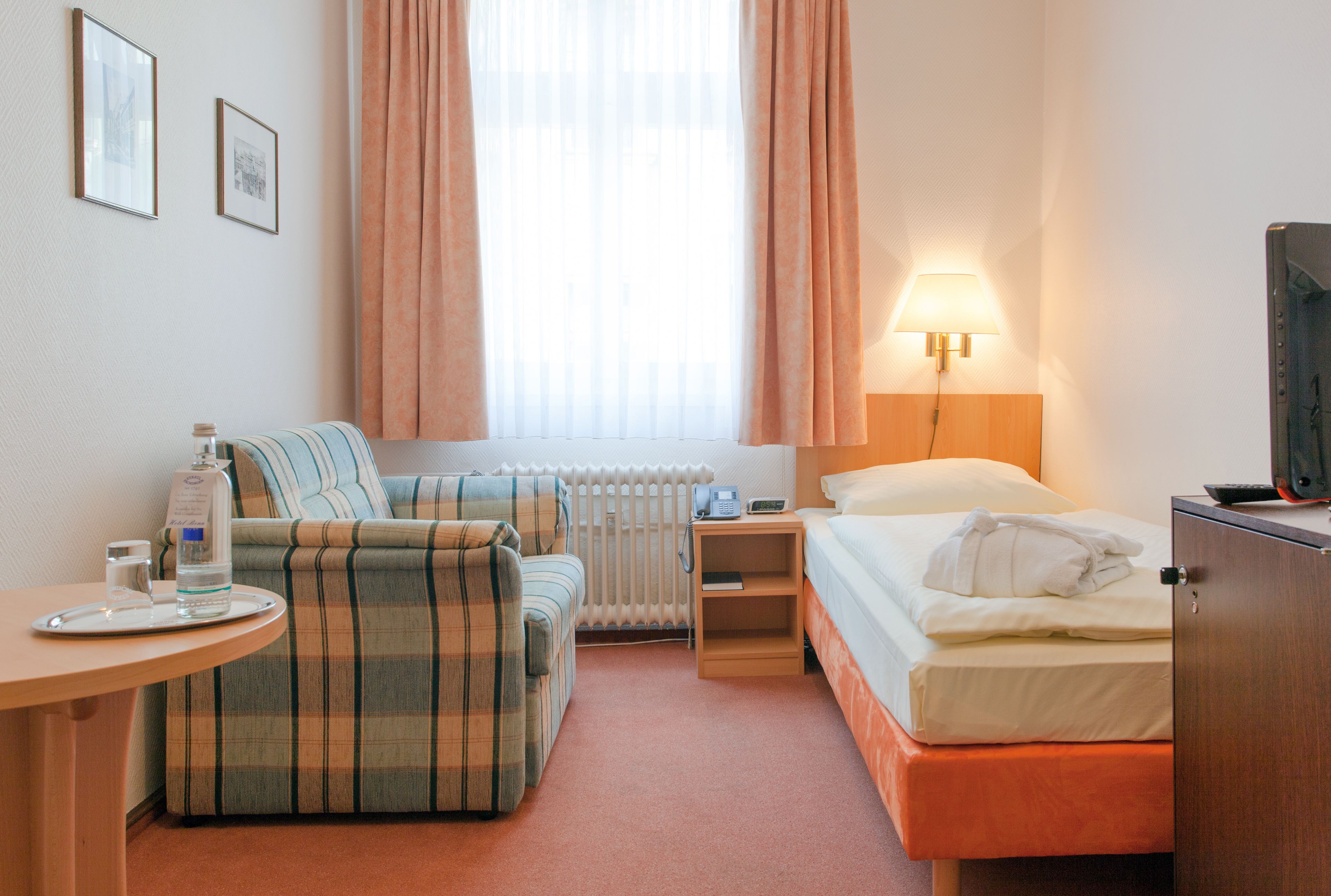 Quarto de hotel com cama, sofá, mesa com um copo de água, luminária e radiador.