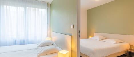 Una habitación de hotel con dos camas, una mesita de noche con lámpara, una ventana con cortinas traslúcidas y un interruptor de luz en la pared.