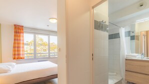 Un dormitorio con cama, ventana con cortinas, y baño con ducha, lavabo y espejo.