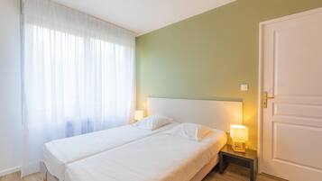 A hotel room with a bed, a nightstand with a lamp, a door, and sheer curtains on the window.